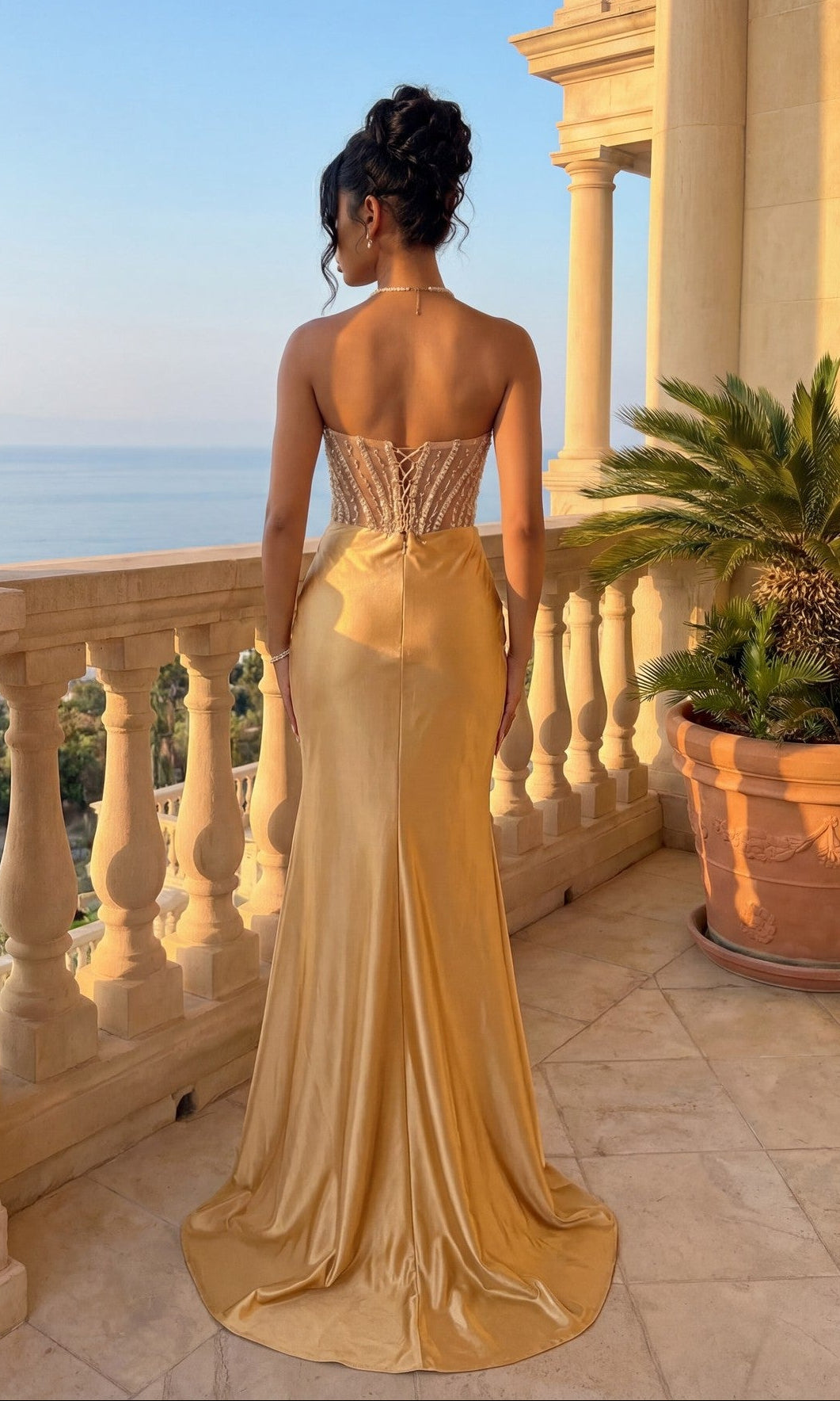A woman in the Amelia Couture 4049 Strapless Long Prom Gown, featuring a gold sweetheart neckline and sheer beaded bodice, stands on a balcony overlooking the sea between columns and a large potted plant.